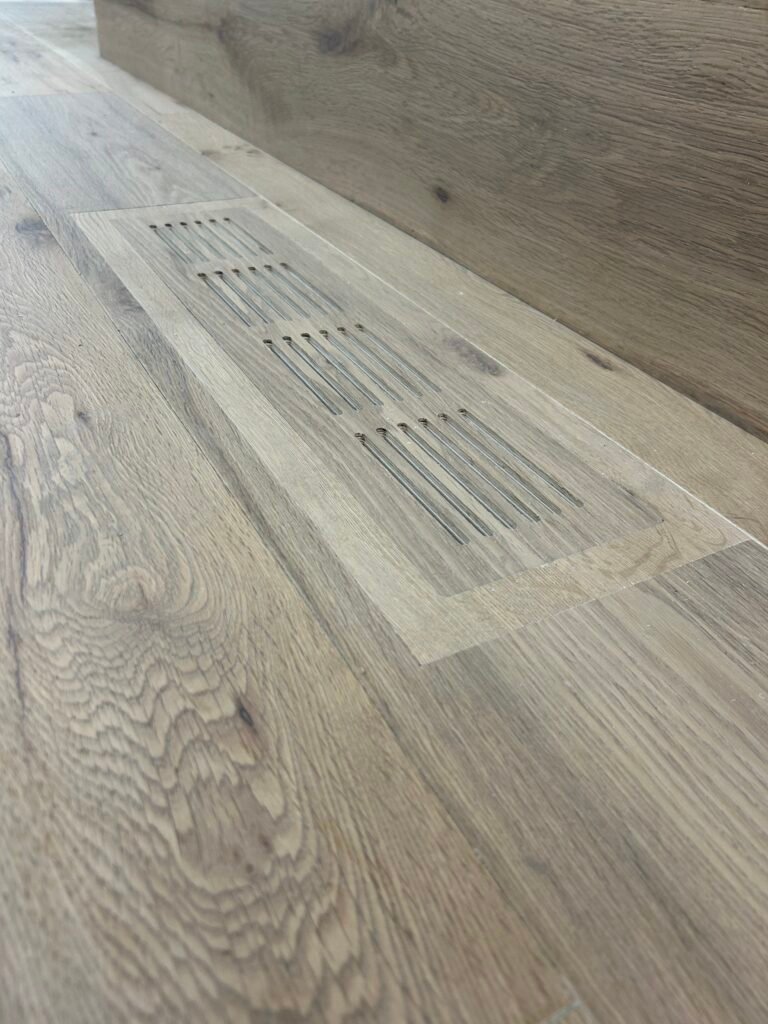 Close-up of a custom oak floor vent cover installed flush in wide-plank hardwood flooring