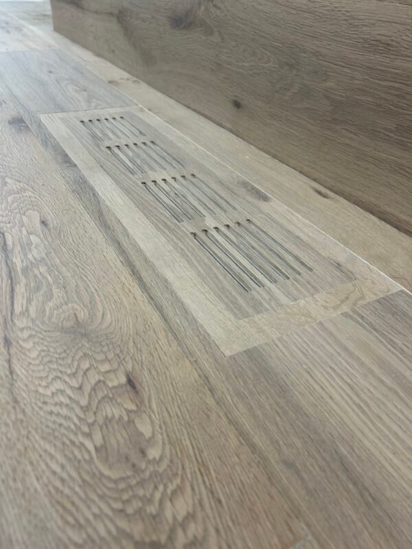 Installed flush vent cover with narrow slots, matching the surrounding wood-look flooring.